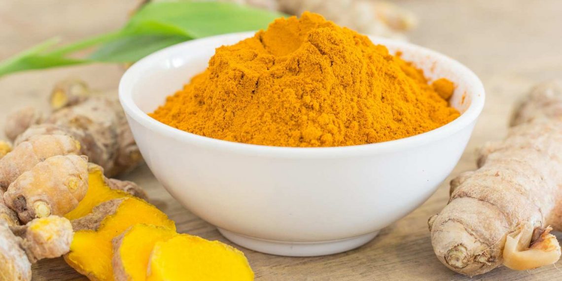 Bowl of turmeric powder with fresh turmeric roots on a wooden table.