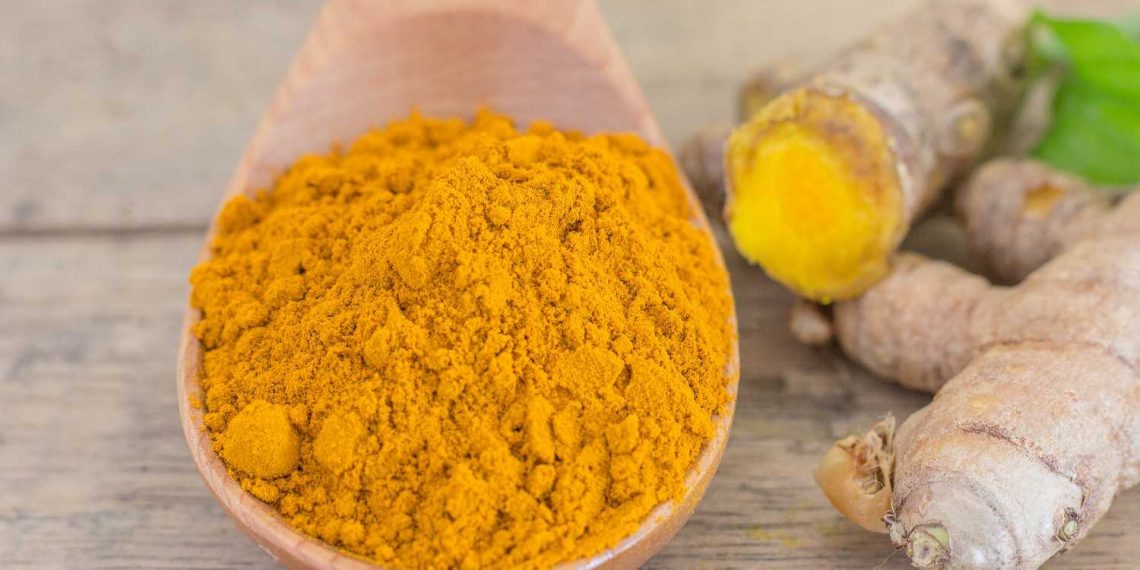 Turmeric powder in a wooden spoon next to fresh turmeric root.