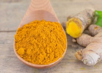 Turmeric powder in a wooden spoon next to fresh turmeric root.