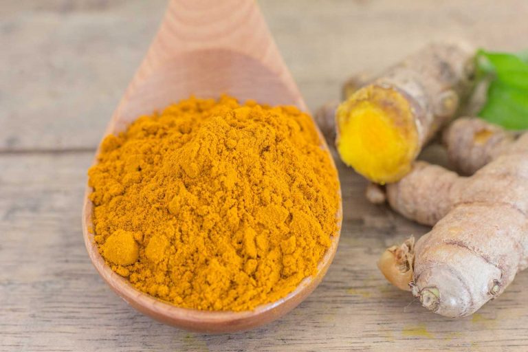 Turmeric powder in a wooden spoon next to fresh turmeric root.