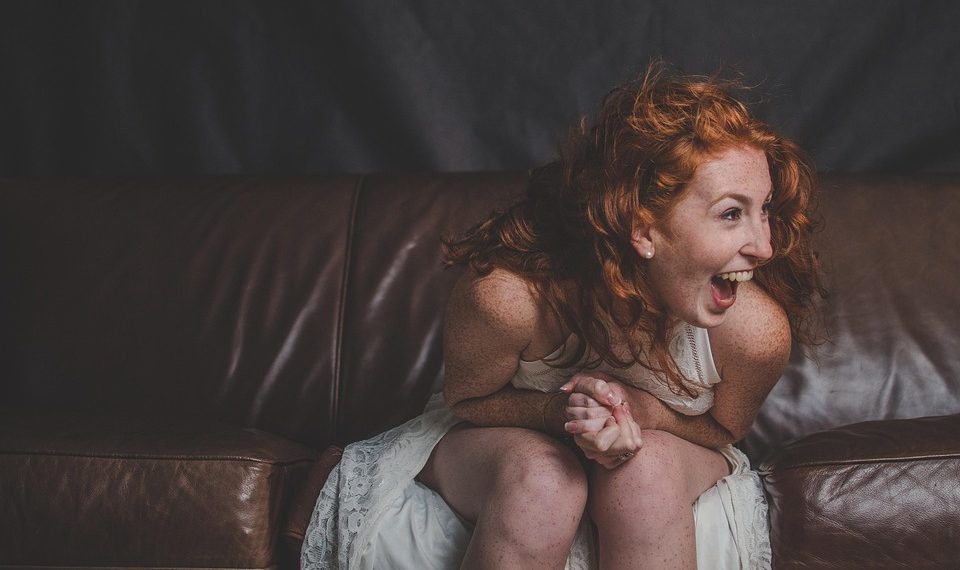 Red-haired woman laughing on a brown leather sofa.