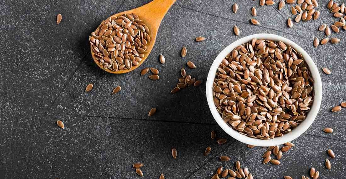 Flaxseeds in a white bowl and wooden spoon on a dark surface.