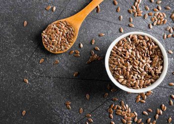 Flaxseeds in a white bowl and wooden spoon on a dark surface.