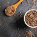 Flaxseeds in a white bowl and wooden spoon on a dark surface.