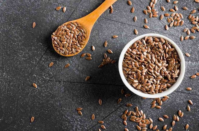 Flaxseeds in a white bowl and wooden spoon on a dark surface.