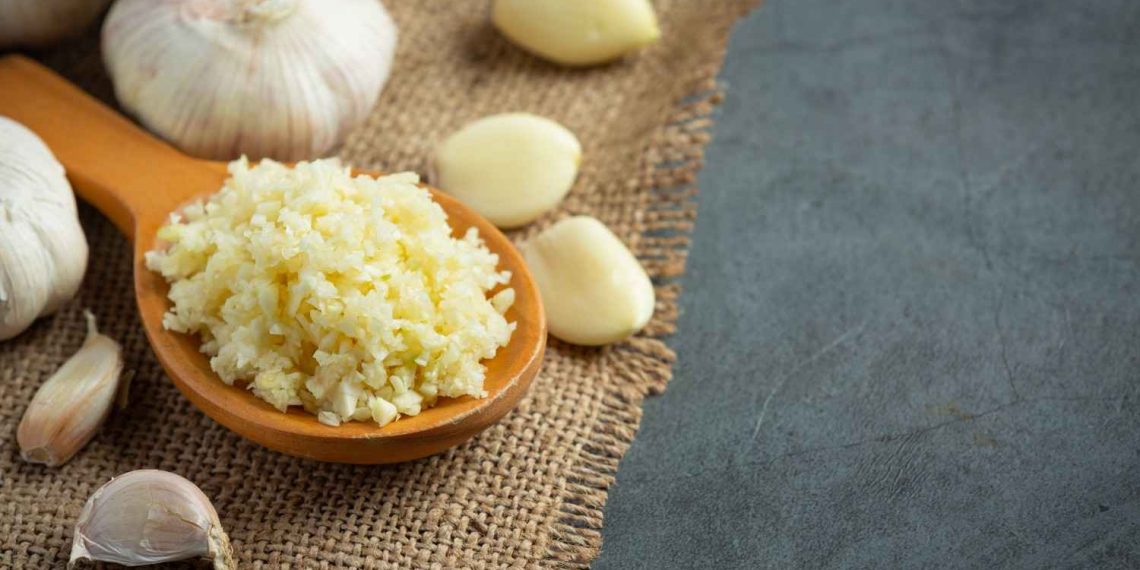 Crushed garlic on a wooden spoon surrounded by cloves on burlap.