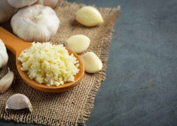 Crushed garlic on a wooden spoon surrounded by cloves on burlap.