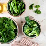 Fresh spinach, peas, and broccoli in bowls on a marble surface.