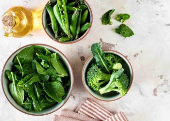 Fresh spinach, peas, and broccoli in bowls on a marble surface.