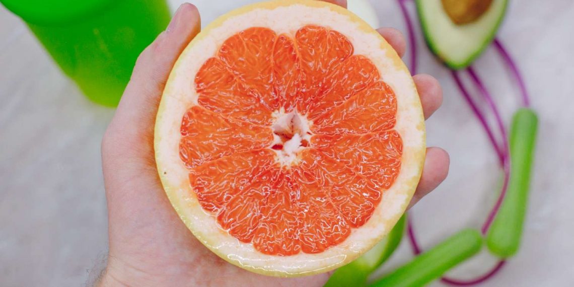 Hand holding a halved grapefruit next to an avocado and a green smoothie.