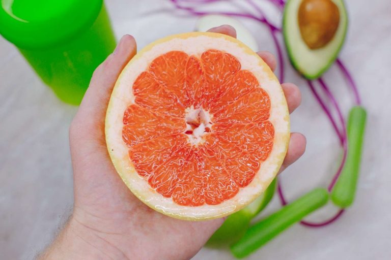 Hand holding a halved grapefruit next to an avocado and a green smoothie.