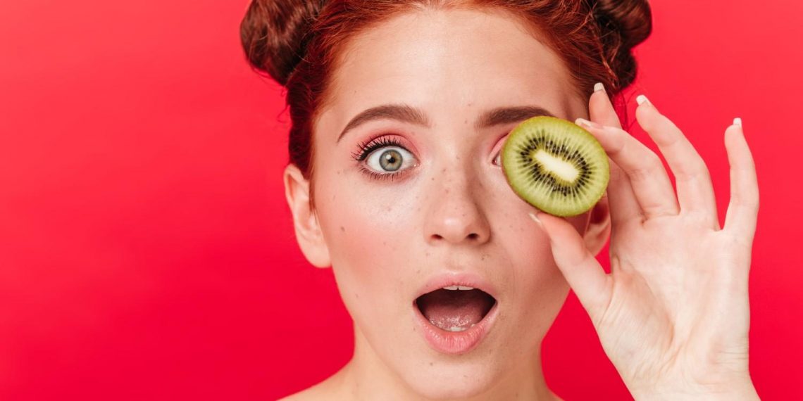 Red-haired woman holding kiwi slice over eye with surprised expression.