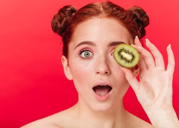 Red-haired woman holding kiwi slice over eye with surprised expression.