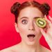 Red-haired woman holding kiwi slice over eye with surprised expression.