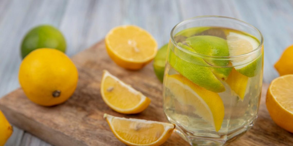 Glass of lemon water with sliced lemons and limes on a wooden board.