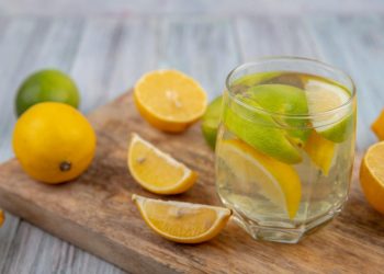 Glass of lemon water with sliced lemons and limes on a wooden board.