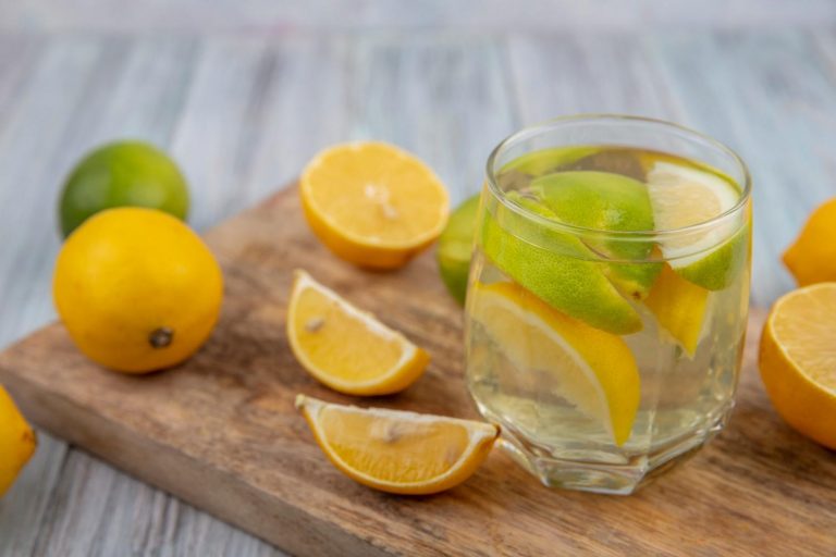 Glass of lemon water with sliced lemons and limes on a wooden board.