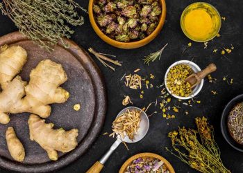 Ginger and herbs on a dark surface with various spices displayed.