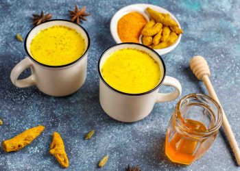 Two mugs of turmeric latte with spices and honey on a blue surface.