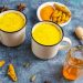 Two mugs of turmeric latte with spices and honey on a blue surface.