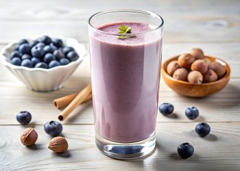 Blueberry smoothie in a glass, garnished with a mint leaf, surrounded by fresh blueberries and macadamia nuts.