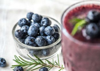 Fresh blueberries in a glass bowl beside a blueberry smoothie garnished with rosemary.