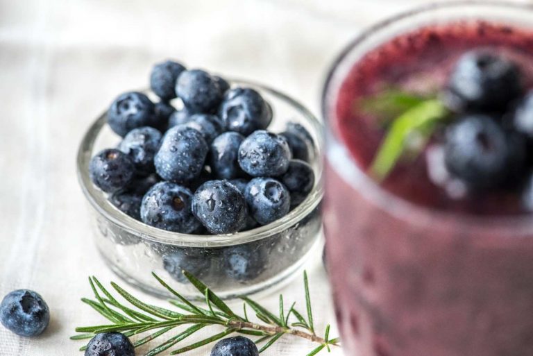 Fresh blueberries in a glass bowl beside a blueberry smoothie garnished with rosemary.