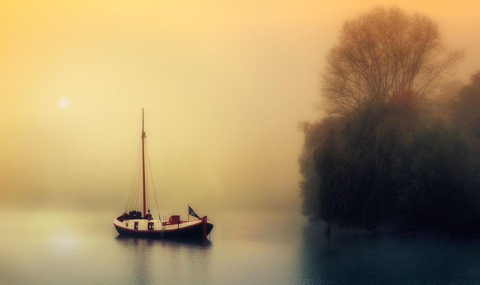 Sailboat on misty lake at sunrise near trees.