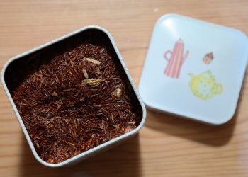 Rooibos tea leaves in an open tin container on a wooden surface.
