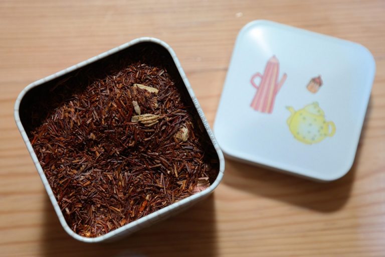 Rooibos tea leaves in an open tin container on a wooden surface.