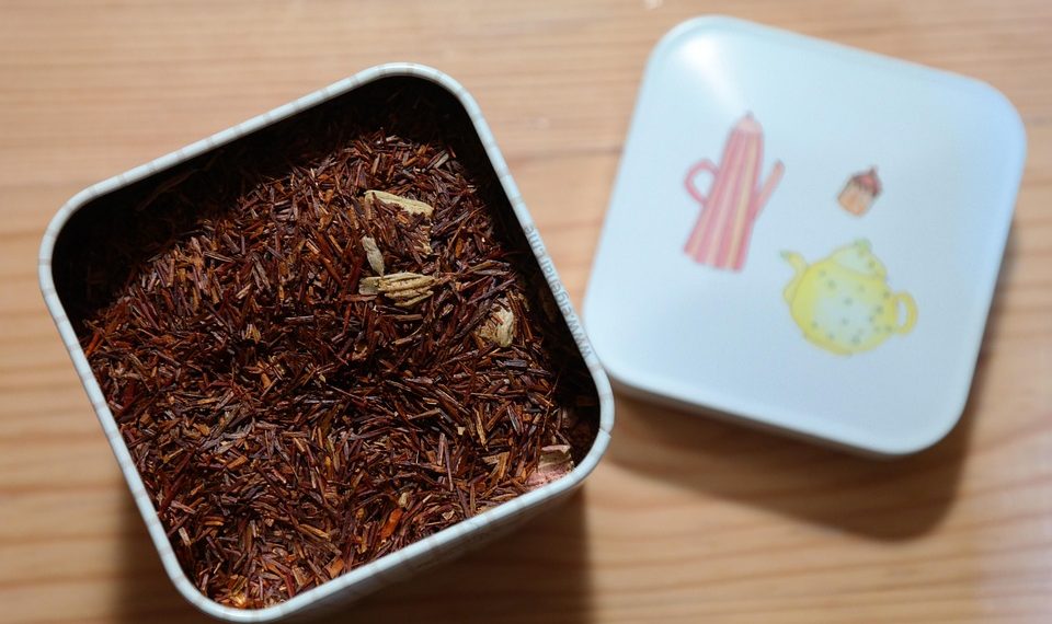 Rooibos tea leaves in an open tin container on a wooden surface.