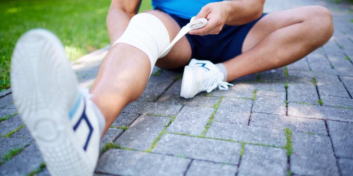 Man wrapping knee with bandage on a park path.