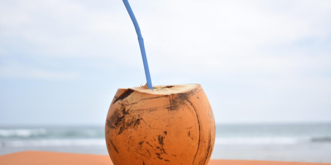 Fresh coconut with straw on sandy beach against ocean backdrop.