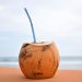 Fresh coconut with straw on sandy beach against ocean backdrop.
