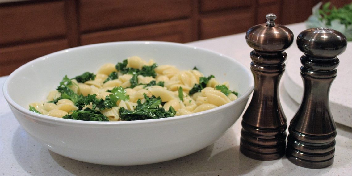 Bowl of pasta with kale next to pepper grinders on a kitchen counter.