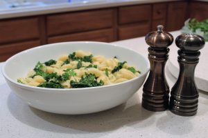 Bowl of pasta with kale next to pepper grinders on a kitchen counter.