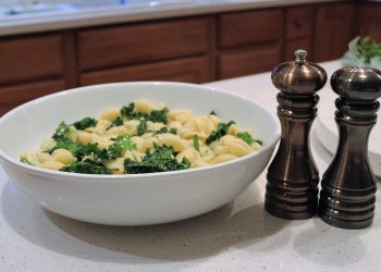 Bowl of pasta with kale next to pepper grinders on a kitchen counter.