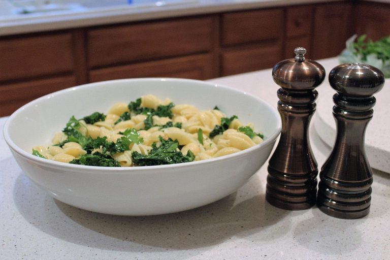 Bowl of pasta with kale next to pepper grinders on a kitchen counter.
