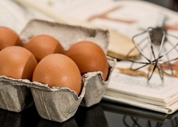 Eggs in carton with a whisk and cookbook in background.