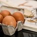 Eggs in carton with a whisk and cookbook in background.