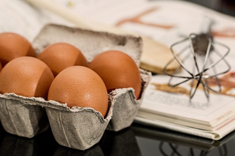 Eggs in carton with a whisk and cookbook in background.