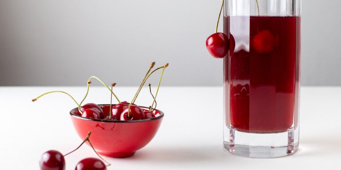 Cherries in red bowl and glass of cherry juice on white table.