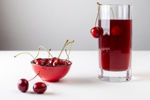 Cherries in red bowl and glass of cherry juice on white table.