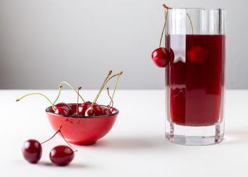 Cherries in red bowl and glass of cherry juice on white table.
