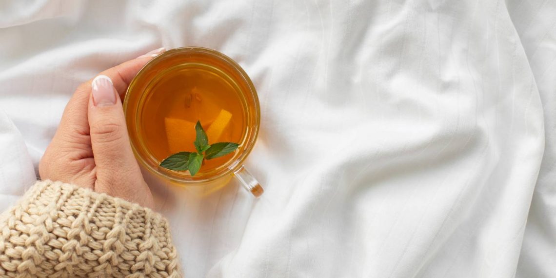Hand holding a cup of herbal tea with mint leaves on a cozy sweater.