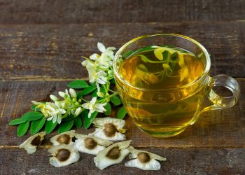 Cup of moringa tea with moringa leaves and seeds on a wooden table.