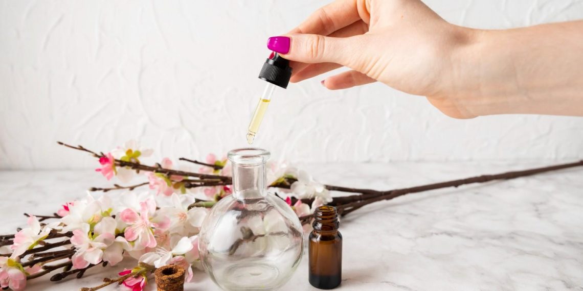 Hand adding oil to a glass bottle with flowers in background.