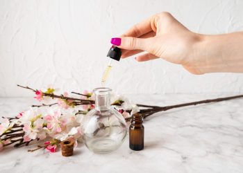 Hand adding oil to a glass bottle with flowers in background.