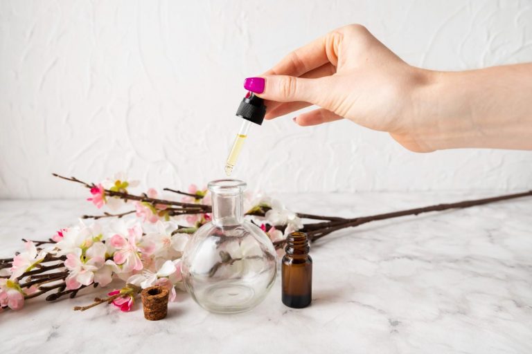 Hand adding oil to a glass bottle with flowers in background.