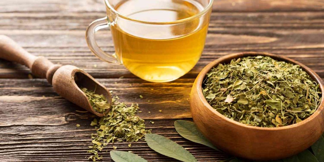 Cup of moringa tea beside a bowl of dried moringa leaves on a wooden table.